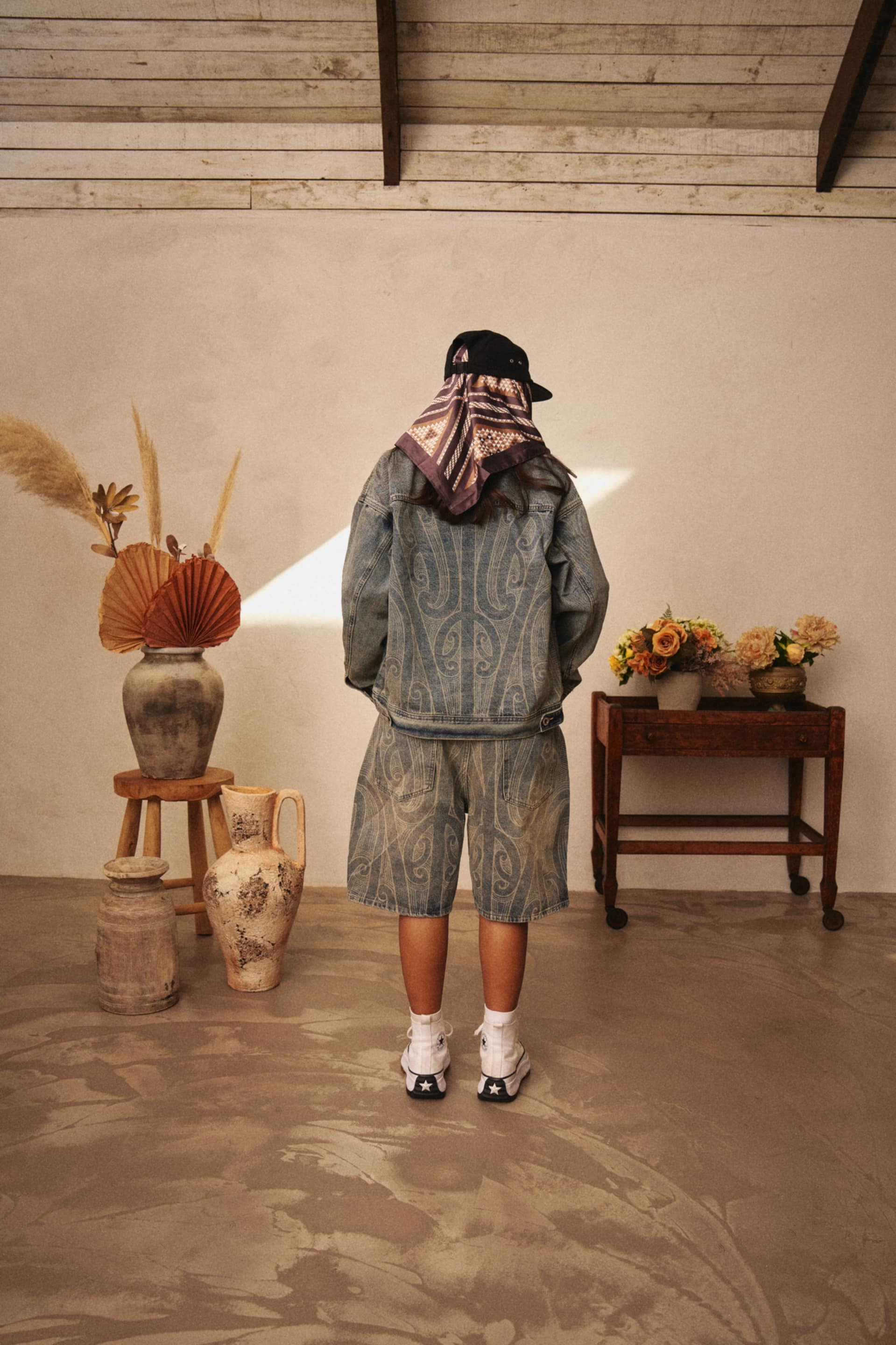 Person wearing L.EYES & CO Denim Jorts and Jacket set and Brown Te Piringa Satin scarf. Māori patterns adorn the scarf and denim clothing.